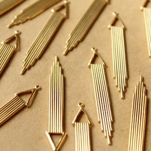 Puede incluir: Una colección de colgantes de metal dorado con un diseño geométrico con múltiples líneas verticales. Los colgantes están dispuestos sobre una superficie marrón.