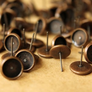 May include: A close-up of a group of antique bronze earring settings. The settings are round and have a small hole in the center for attaching an earring post. The settings are scattered on a brown surface.