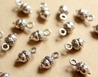 May include: Silver-colored acorn charms with a loop for jewelry making. The charms have detailed caps and smooth bodies. The acorns are scattered on a light brown surface.