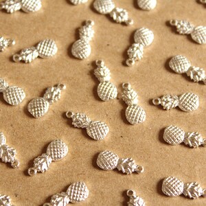May include: A collection of silver pineapple charms, scattered on a brown surface. The charms are detailed with textured pineapple skin and leaves.