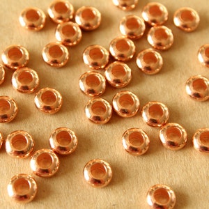 May include: A scattering of small, copper-coloured beads. Each bead has a rounded shape with a hole through the centre. The beads are spread across a light brown surface, reflecting light. These beads are suitable for jewellery making and crafts.