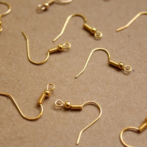 May include: A close-up of a group of gold earring hooks. The hooks are made of metal and have a small loop at the top. They are arranged in a random pattern on a brown surface.