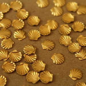May include: A collection of small, gold-colored seashell charms. Each charm is intricately detailed with a fan-like pattern, resembling a miniature scallop shell. The charms are scattered across a neutral surface, creating a textured and decorative effect.
