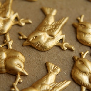 Könnte beinhalten: Nahaufnahme mehrerer goldfarbener Vogelverzierungen. Jeder Vogel sitzt auf einem kleinen Ast. Die Vögel sind detailliert mit Flügeln, Schnäbeln und Augen. Der Hintergrund ist eine neutrale, beige Farbe.