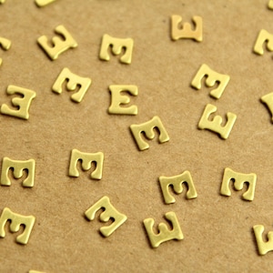 May include: A collection of small, gold-colored metal letter "E" charms scattered on a brown surface. The charms are uniformly shaped and appear to be for jewelry making or crafting.
