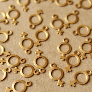 May include: A scattering of small, gold-coloured metal charms, each shaped like the female gender symbol. These charms feature a circular centre, a cross-like shape below, and a small loop at the top for attaching to jewellery or crafts.