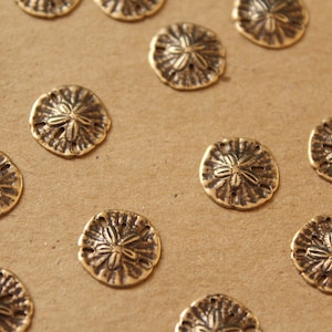 May include: A collection of 14 gold-toned metal sand dollar charms. The charms are arranged in a scattered pattern on a brown surface.