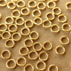 May include: A large quantity of small, gold-colored jump rings. The rings are scattered on a brown surface.