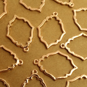 May include: Close-up of several gold-colored metal filigree charms. Each charm has a decorative, openwork design with a scalloped edge and a small loop at the top for attaching to other components. The charms are scattered on a gold background.