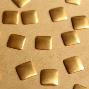 May include: Close-up of several small, square, gold-colored metal embellishments scattered on a light brown surface. The embellishments have rounded corners and a slightly textured appearance, suggesting they are decorative elements.