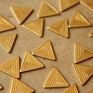 Könnte beinhalten: Eine Sammlung goldfarbener Metalldreiecke mit geometrischem Muster. Die Dreiecke sind auf einer braunen Oberfläche verstreut.