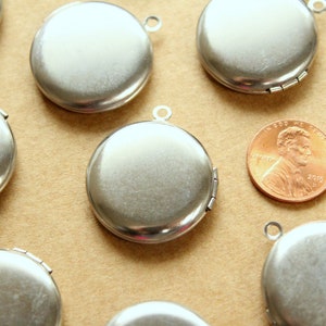 May include: A group of eight silver locket pendants with a hinged opening. The lockets are round and have a smooth, shiny surface. They are arranged on a brown surface.