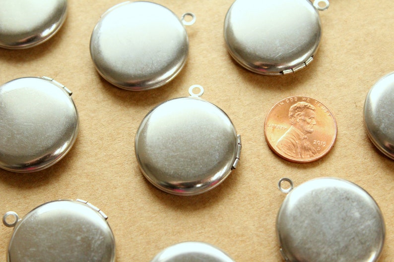 May include: A group of eight silver locket pendants with a hinged opening. The lockets are round and have a smooth, shiny surface. They are arranged on a brown surface.