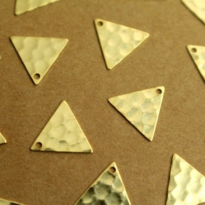 May include: Close-up of several gold-colored, hammered-texture triangle charms. Each charm has a small hole at one or two corners, suggesting they are designed for jewelry making or crafting. The charms are scattered on a brown surface.