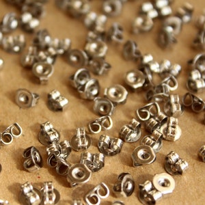 May include: A close-up of a large quantity of silver earring backs. The backs are made of metal and have a small loop on the back for attaching to an earring.
