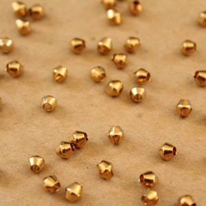 May include: A scattering of small, gold-colored metal beads with a faceted, cone-like shape. The beads are scattered on a light brown surface, creating a textured and reflective display. Ideal for jewelry making and crafts.