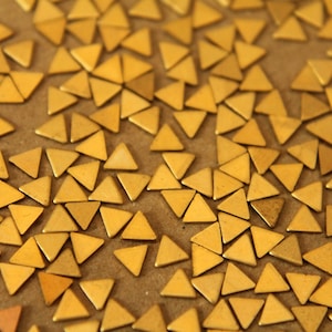 May include: A close-up image of a large quantity of small, gold-coloured, triangular-shaped metal pieces scattered on a brown surface.