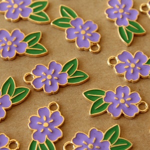 May include: Close-up of several purple flower charms with green leaves and gold-colored edges. Each charm has a small loop for attaching to jewelry or other crafts. The charms are scattered on a light brown surface.