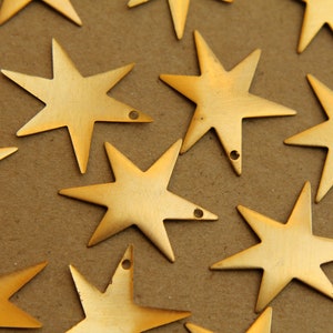 Puede incluir: Una colección de pequeños dijes dorados en forma de estrella. Cada dije tiene siete puntas y un pequeño agujero para ensartar. Los dijes están dispersos sobre una superficie marrón, creando un tema celestial.