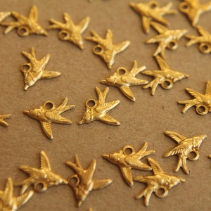 May include: A collection of gold-toned metal bird charms, each with a small loop for attaching to jewelry or other crafts. The charms are scattered on a brown surface.