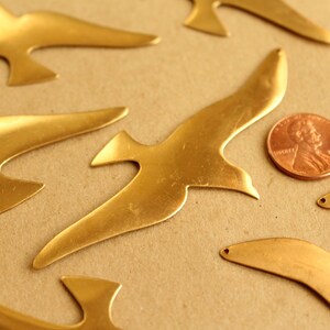 May include: A collection of seven brass bird cutouts in various sizes and shapes. The cutouts are arranged on a brown surface with a penny for scale.