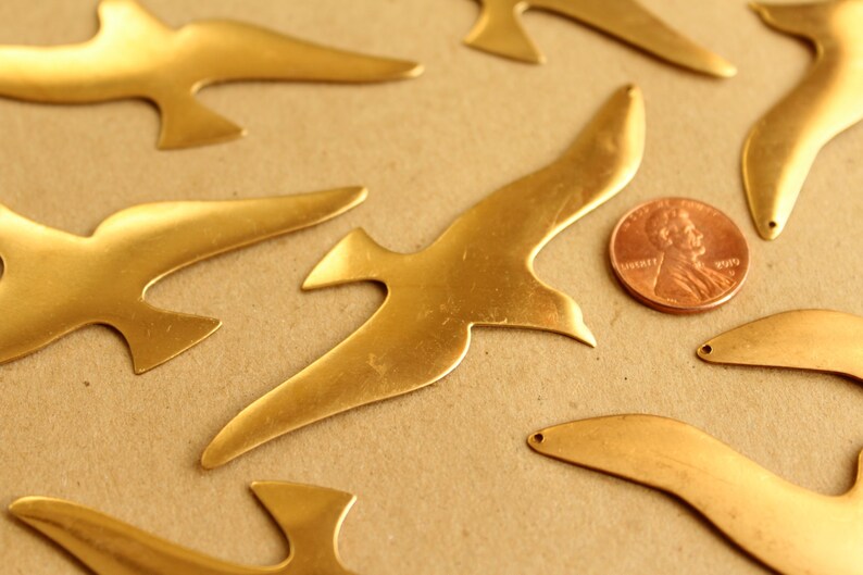 May include: A collection of seven brass bird cutouts in various sizes and shapes. The cutouts are arranged on a brown surface with a penny for scale.
