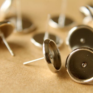 May include: A close-up of silver earring settings with a round, flat base and a small hole in the center. The settings are scattered on a brown surface.