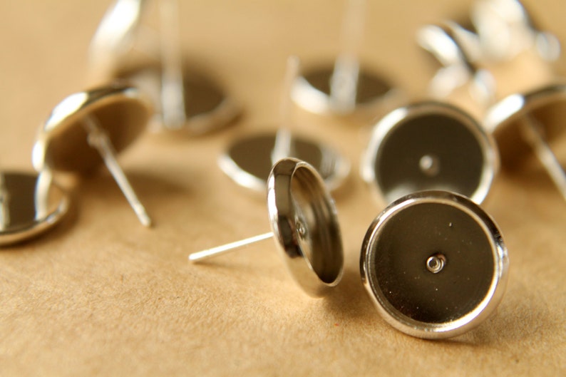 May include: A close-up of silver earring settings with a round, flat base and a small hole in the center. The settings are scattered on a brown surface.