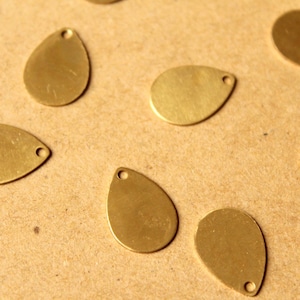 May include: A collection of gold-colored metal teardrop-shaped charms with a hole at the top. The charms are arranged on a brown surface.