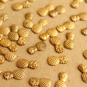 May include: A close-up of a large quantity of gold pineapple-shaped metal charms. The charms are scattered on a brown surface.