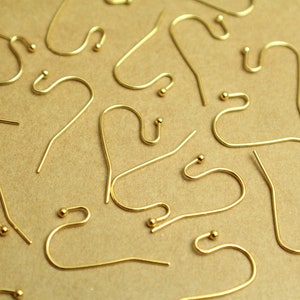 Könnte beinhalten: Nahaufnahme einer Gruppe von goldenen Ohrhaken, die auf einer braunen Oberfläche verstreut sind. Die Haken sind aus Metall und haben eine kleine Kugel am Ende.