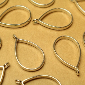 May include: A group of silver teardrop-shaped pendant blanks, with a loop at the top for attaching a chain or cord. The blanks are arranged on a brown surface.