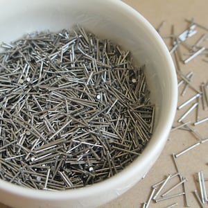 May include: A white ceramic bowl filled with a large quantity of silver-colored straight pins. Some pins are scattered on a brown surface.