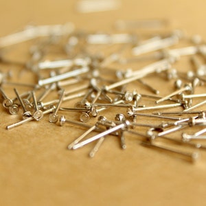 May include: A close-up of a pile of silver-toned metal pins with round heads. The pins are scattered on a brown surface.