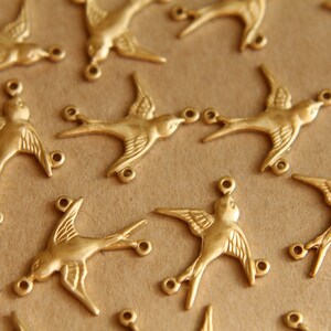 May include: A collection of gold-toned metal bird charms with open jump rings. The charms are in the shape of a stylized bird with wings outstretched.