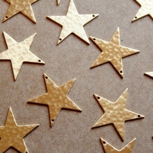 May include: A collection of gold-toned metal star shapes with a hammered texture. The stars have five points and are scattered on a brown surface.