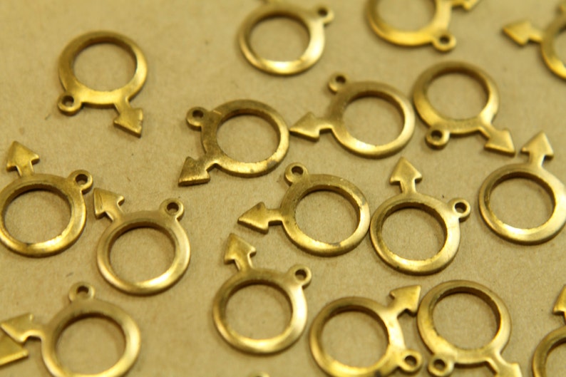 May include: A group of gold-colored metal charms in the shape of a circle with an arrow pointing to the right. The charms are scattered on a brown surface.