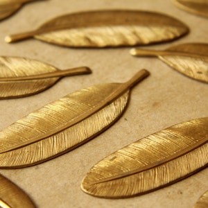 May include: A collection of gold-colored metal feather-shaped ornaments. The feathers have a textured surface and are arranged on a brown background.
