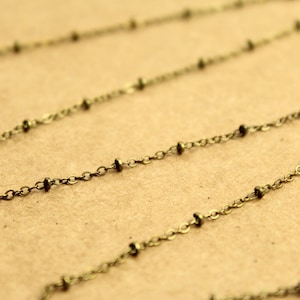 May include: Close-up of a delicate antique brass chain with small, evenly spaced beads. The chain is displayed against a neutral tan background, highlighting its intricate design and vintage aesthetic. Ideal for jewelry making.