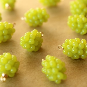 May include: Close-up of several green grape-shaped beads with gold-colored loops. The beads are a light, translucent green color and appear to be made of plastic or resin. These beads are likely used for jewelry making or other craft projects.