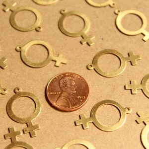May include: A close-up of a group of gold-colored metal charms shaped like the female symbol. The charms are scattered on a brown surface, with a penny in the center. The penny is dated 2012 and has the words "In God We Trust" and "Liberty" inscribed on it.