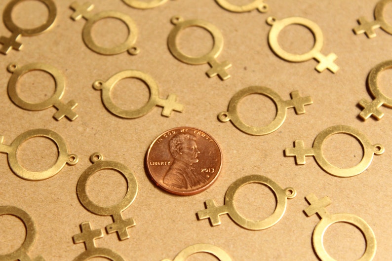 May include: A close-up of a group of gold-colored metal charms shaped like the female symbol. The charms are scattered on a brown surface, with a penny in the center. The penny is dated 2012 and has the words "In God We Trust" and "Liberty" inscribed on it.
