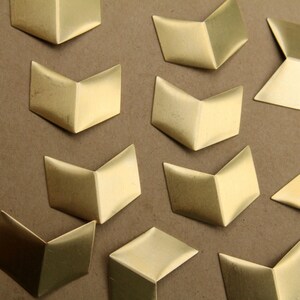 May include: A collection of gold-coloured metal corner brackets in various shapes, including arrow and square designs. The brackets are arranged on a brown surface.