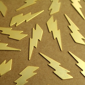 May include: A close-up of a group of gold lightning bolt shaped metal charms. The charms are scattered on a brown surface.
