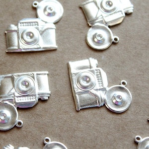 May include: A set of silver-toned camera charms with a lens cap charm. The charms are detailed with a vintage camera design.