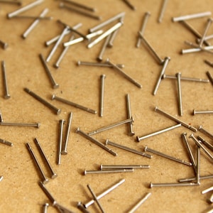 May include: A collection of small, silver-colored metal pins scattered on a light brown surface. The pins have a cylindrical body with a flat head. The image is a close-up shot, highlighting the details of the pins and the texture of the surface.