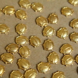 Puede incluir: Una colección de abalorios en forma de escarabajo de color dorado, esparcidos sobre una superficie marrón. Los abalorios son detallados y tienen un acabado texturizado.