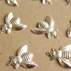 Puede incluir: Una colecci&oacute;n de abalorios de abeja plateados con alas y cuerpos texturizados. Los abalorios est&aacute;n dispuestos sobre una superficie marr&oacute;n.