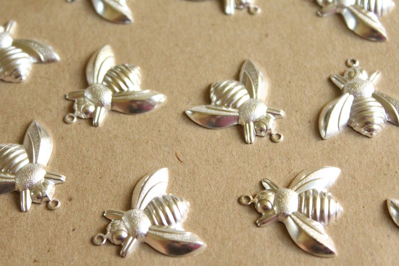 Puede incluir: Una colecci&oacute;n de abalorios de abeja plateados con alas y cuerpos texturizados. Los abalorios est&aacute;n dispuestos sobre una superficie marr&oacute;n.