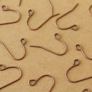 May include: Close-up of several copper-colored earring hooks. The hooks are arranged on a light brown surface. Each hook has a curved design with a small loop at the top and a straight wire extending from the bottom.